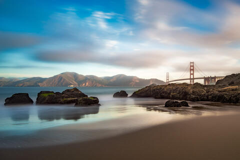 Baker Beach in San Francisco with smooth ocean water, rocks in the foreground, and Golden Gate Bridge in the distance