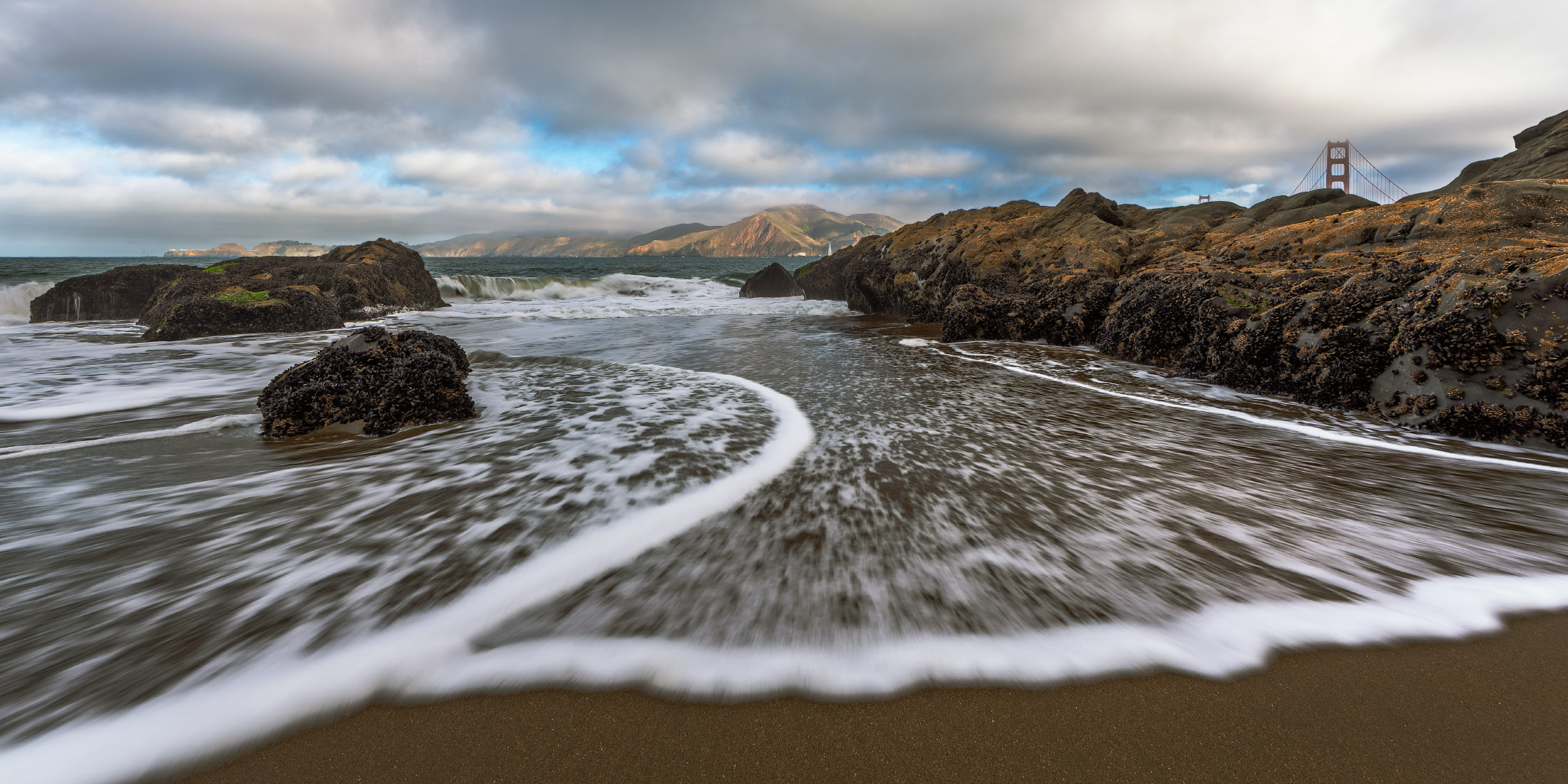 Cold Rush Baker Beach in San Francisco with waves along the shore and partial view of the Golden Gate Bridge
